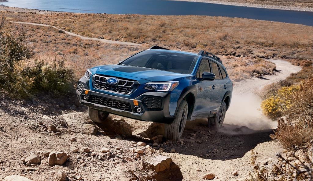 Subaru Outback Wilderness 2025 bleu grimpe une pente rocheuse dans un décor désertique avec un lac en arrière-plan.