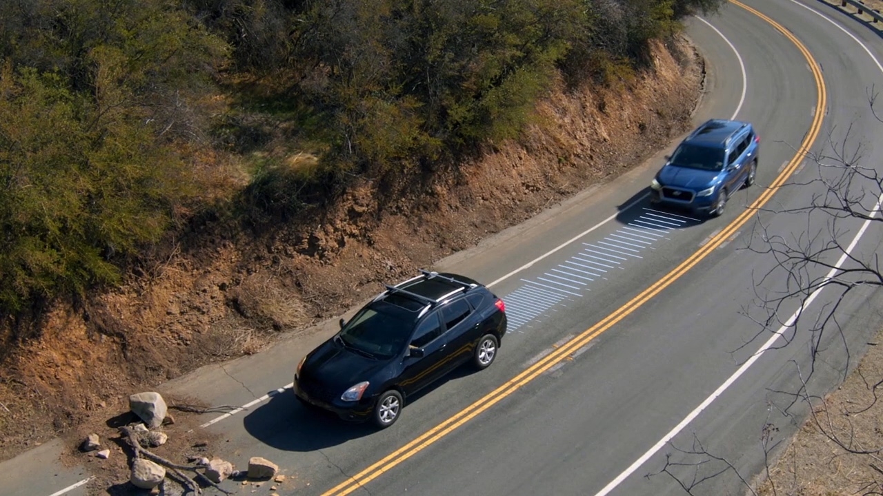 Vue aérienne d’un Subaru bleu activant le freinage précollision devant un VUS noir immobilisé sur une route de montagne avec débris au sol.